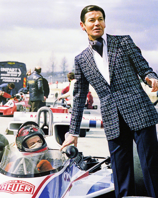 Man in a suit standing next to a Formula 1 car with a driver inside, on a race track.