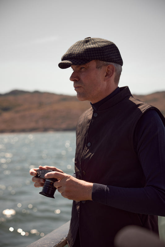A man in profile wearing a dark patterned flat cap, a dark quilted vest over a long-sleeve shirt, and holding binoculars near a body of water with hills in the background.