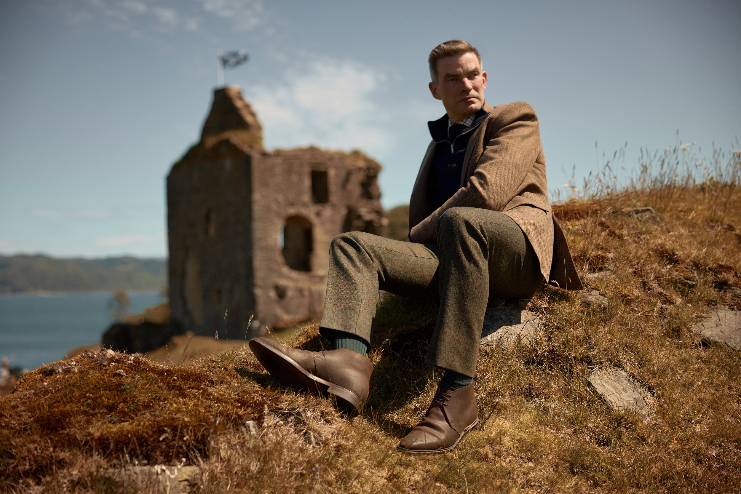 A man wearing a brown tweed blazer, olive green trousers, and brown leather shoes sits on a grassy hillside with a stone ruin and water in the background.