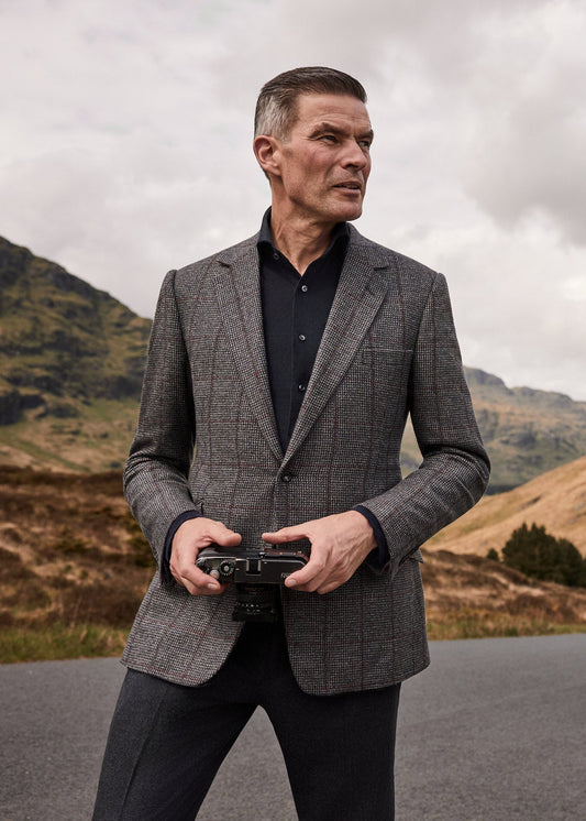 A man in a gray patterned blazer and black shirt stands on a road holding a camera, with a black Aston Martin car parked on the road behind him against a backdrop of brown hills and a cloudy sky.