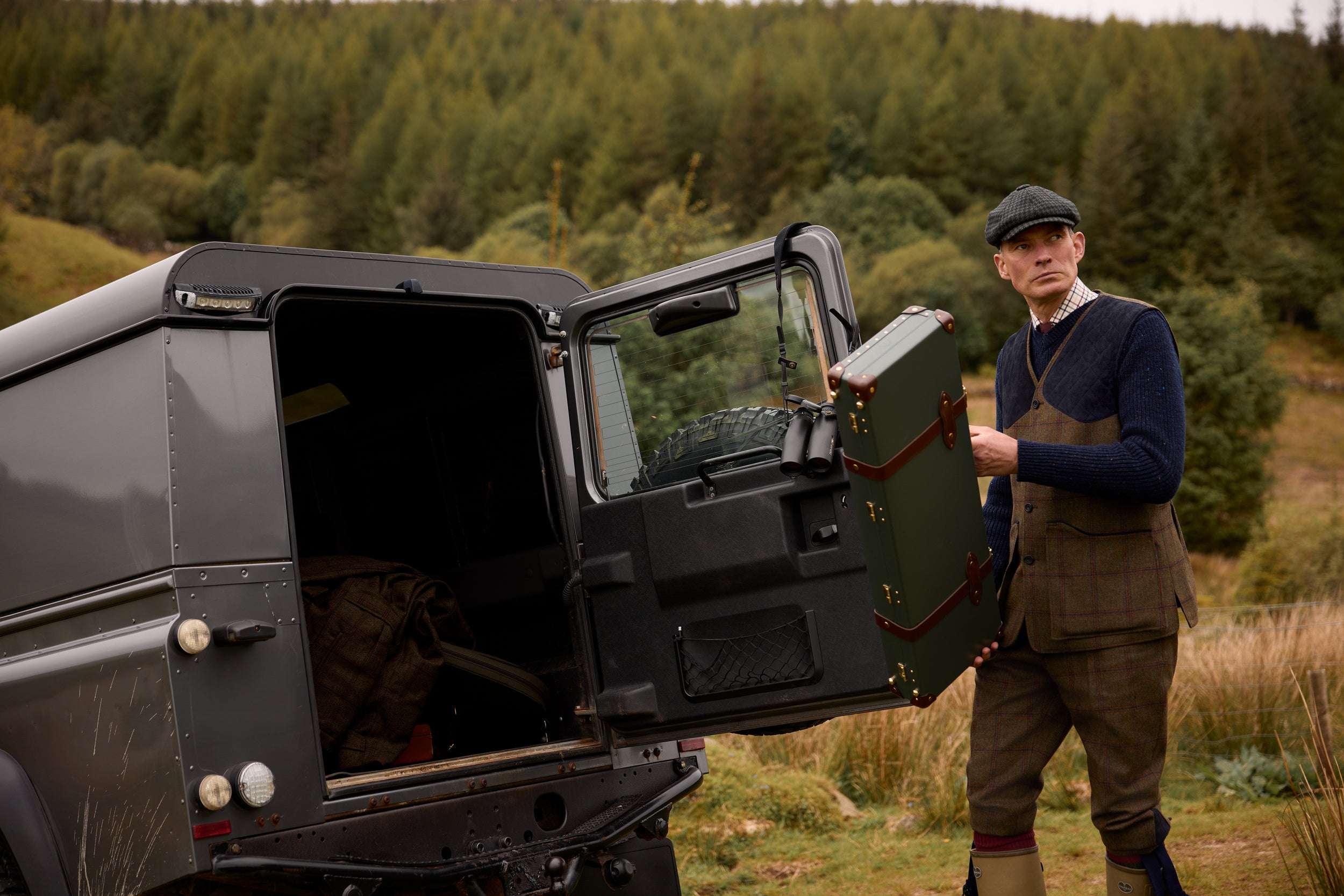 A man in traditional hunting attire stands next to a dark gray off-road vehicle with its rear door open, holding a green hard-sided storage case in an outdoor grassy landscape with a forest in the background.